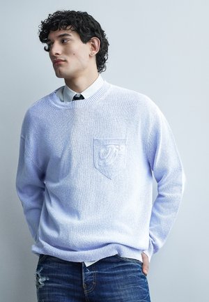 Jeune homme aux cheveux bouclés et foncés, portant un pull bleu clair par-dessus une chemise blanche et une cravate noire, debout les mains dans les poches, regardant vers la gauche.