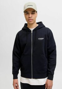 Navy zip-up hoodie with drawstrings and front pockets, featuring white logo on left chest. Cap in light beige with black text.