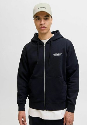 Navy zip-up hoodie met trekkoorden en voorzakken, met een wit logo op de linkerborst. Pet in lichtbeige met zwarte tekst.