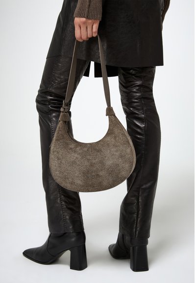 Borsa rotonda e morbida in pelle taupe con una superficie texturizzata e una sottile tracolla regolabile. Indossata all'altezza del braccio con pantaloni in pelle scuri e stivali alla caviglia.