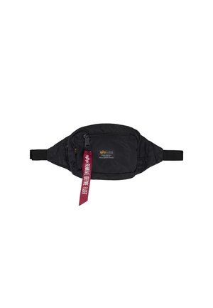 Sac banane noir avec poches zippées et une étiquette en tissu rouge indiquant "Remove Before Flight", attachée à la fermeture éclair avant.