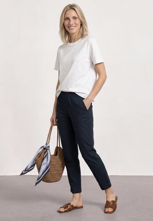 Femme souriante aux cheveux blonds portant un t-shirt blanc, un pantalon noir, des sandales marron, tenant un sac tissé avec un foulard noué.