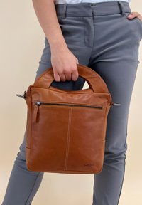 Brązowa skórzana torebka na ramię z podwójnymi uchwytami, zamykaną na zamek przednią kieszenią, gładką teksturą i kontrastowymi detalami w szwie. Wymiary są kompaktowe i pionowe.