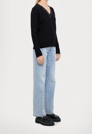 Femme portant un pull noir col en V avec des détails multicolores sur les bords, un jean droit bleu clair, et des chaussures noires à plateforme épaisse, debout de profil.