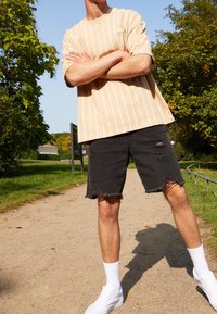 Beige raidoitettu t-paita, rento malli, yhdistetty mustiin kuluneisiin shortseihin. Valkoiset lenkkarit, nilkkasukat, ulkoilmaympäristö, jossa on vihreää kasvillisuutta.