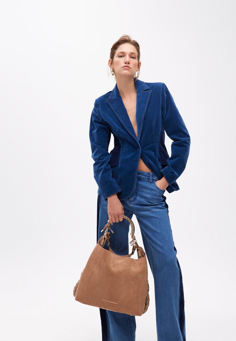 Una modella posa con sicurezza indossando un blazer di velluto blu e jeans blu, mentre tiene una borsa in corduroy beige contro uno sfondo bianco semplice.