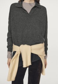 Maglione lavorato a maglia color carbone con scollo con colletto, indossato sopra una maglietta grigio chiaro, con un maglione color crema annodato in vita.