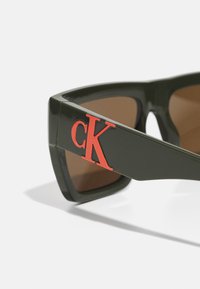 Gröna överdimensionerade solglasögon med bruna linser, som har en framträdande röd "cK"-logotyp på sidan. Glansig yta, vinklad design.
