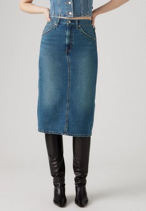 Kvinde iført en blå denim nederdel til knæet og sorte knæhøje støvler, med hænderne hvilende på hofterne mod en ensfarvet baggrund.