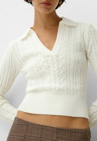 Biały sweter z dzianiny warkoczowej z dekoltem w serek i kołnierzykiem. Posiada ściągacz na dole i teksturowany wzór; dopasowany krój.