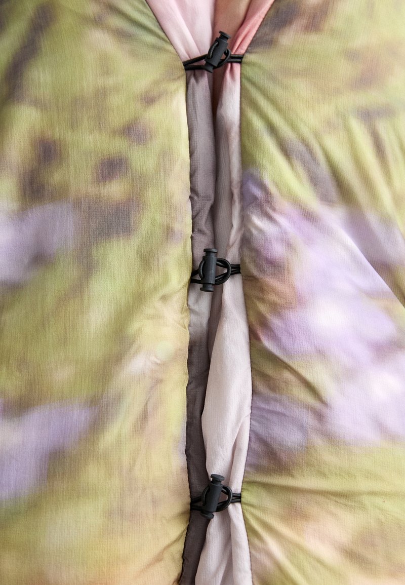 Barevná látka s rozmazaným květinovým vzorem v odstínech zelené, růžové a fialové, upevněná černými plastovými přezkami.