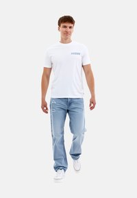 Camiseta blanca de manga corta con un gráfico azul en el pecho, combinada con jeans desgastados de color azul claro y zapatillas blancas.
