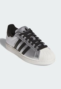 Baskets argentées avec accents noirs, présentant trois bandes noires, une coque avant blanche texturée et des lacets noirs. Matériau lisse et col rembourré.