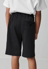Schwarze Shorts mit glatter Textur, elastischem Bund und einer einzigen Gesäßtasche mit Knopfdetail. Kombination mit einem weißen Hemd.