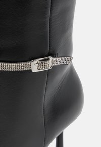 Botte en cuir noir avec une boucle en argent brillant et une sangle ornée de strass. La botte a une texture lisse et une pointe effilée.