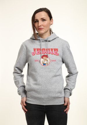 Frau trägt grauen Kapuzenpullover mit dem Text "JESSIE EST. 1999" und einer Cartoon-Cowgirl-Figur auf der Vorderseite, steht mit Händen in der Nähe der Taschen.