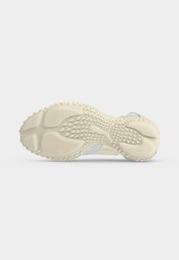 Semelle d'une chaussure de sport beige clair avec des motifs de grip texturés et un logo en relief au centre, présentée sur fond blanc.