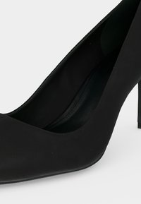 Chaussure pour femme noire à talon haut avec un bout pointu et une finition mate lisse sur fond blanc.