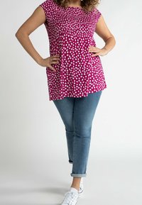 Mujer que lleva una blusa sin mangas de flores magenta, jeans azules enrollados y zapatillas blancas, con las manos en las caderas frente a un fondo sencillo.