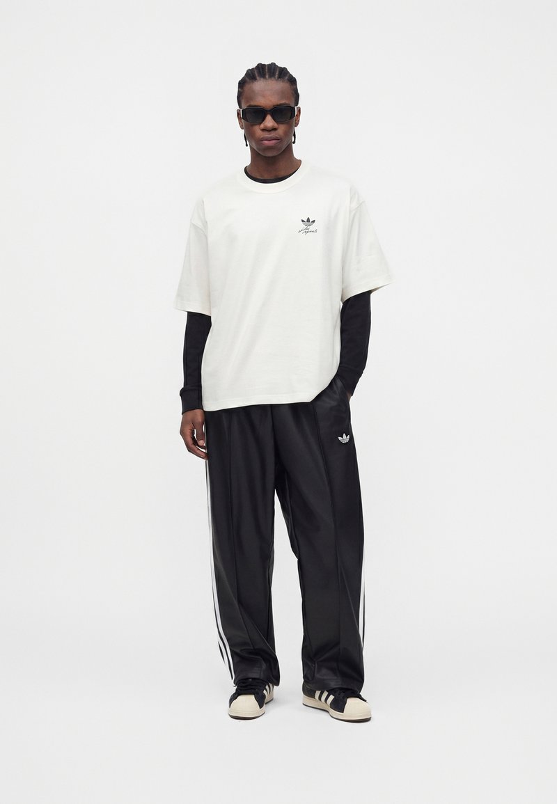 Homme aux cheveux tressés portant des lunettes de soleil noires, un t-shirt blanc oversize par-dessus des manches longues noires, un pantalon de survêtement noir et des baskets noir et blanc.