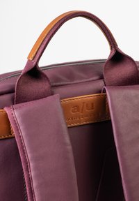 Zaino in tessuto viola con dettagli in pelle marrone, dotato di maniglia superiore e spallacci imbottiti. Il logo del marchio è impresso su un'etichetta in pelle.