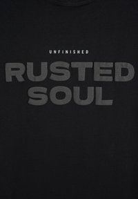 Czarny T-shirt z wyrazistym, szarym napisem. U góry mniejszą czcionką znajduje się słowo "UNFINISHED", poniżej większymi, łagodniejszymi literami napis "RUSTED SOUL".