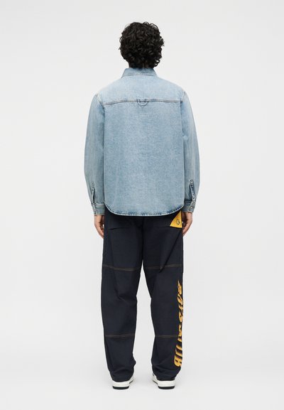 Άτομο με σγουρά μαλλιά που φοράει ανοιχτό μπλε τζιν μπουφάν και μαύρο παντελόνι με μεγάλο κίτρινο κείμενο στο δεξί πόδι, στέκεται γυρισμένο προς τα πίσω.