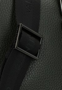 Sangle en nylon noir avec boucle carrée en métal noir, attachée à une surface en cuir vert foncé texturé, avec un logo en relief.