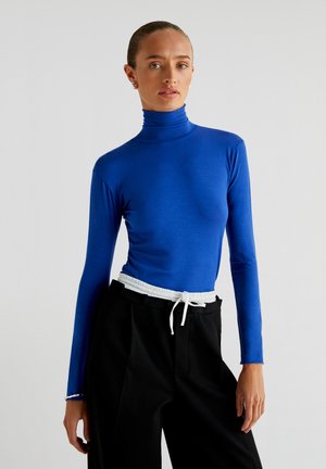 Sinine pika varrukaga turtleneck, mis on valmistatud siledast kangast, koos kõrge vöökohaga mustade pükstega, millel on valge paelavöö.