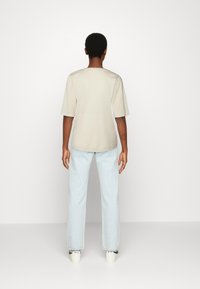 Beige kortærmet skjorte med rund hals, parret med lyseblå lige jeans og hvide sneakers, set bagfra.