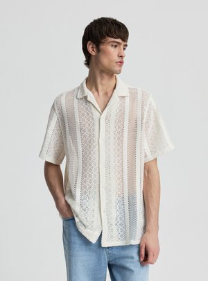 Giovane uomo che indossa una camicia bianca a maniche corte, lavorata a maglia aperta con bottoni e jeans azzurri, guardando di lato su uno sfondo semplice.