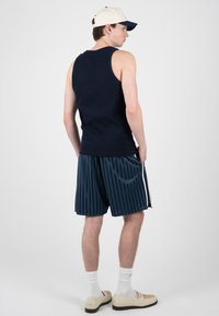 Homme portant une casquette beige, un débardeur noir, un short rayé bleu, des chaussettes blanches et des mocassins beiges, debout, de dos.