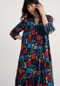 Sukienka maxi w kolorowym wzorze patchworkowym, z kwadratami w kwiatowe desenie. Materiał wydaje się lekki, w luźnym kroju i z dekoltem w kształcie V.