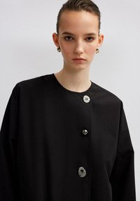 Čierna oversized bunda s kruhovým výstrihom, so striebornými a škvrnitými gombíkmi, hladkou textúrou a širokými rukávmi.