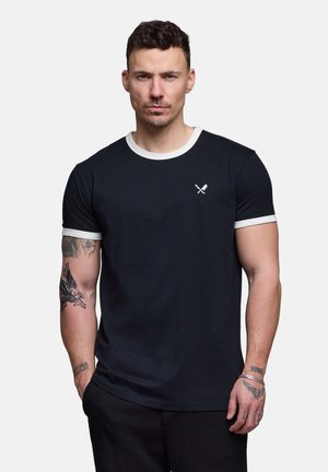 Mann med kort mørkt hår iført svart T-skjorte med hvit kant og liten logo med krysset kniv og gaffel, stående med hendene delvis i lommene.