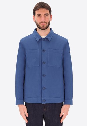 Uomo di fronte che indossa una giacca blu con bottoni e due tasche sul petto e jeans scuri su uno sfondo semplice.