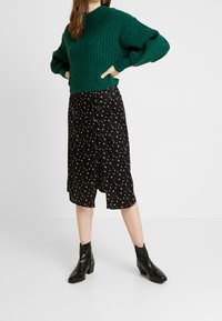 Zelený žebrovaný svetr s nadýchanými rukávy, kombinovaný s černou sukní s květinovým vzorem a černými kotníkovými botami s masivním podpatkem.