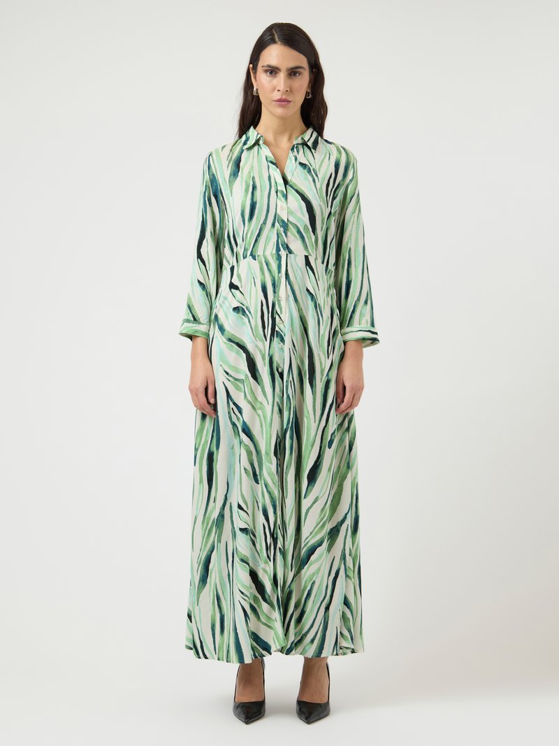 Naine seisab, kandes pikka rohelist ja valget abstraktset mustriga kleiti, millel on pikad varrukad, ning musti kõrgeid kontsasid.
