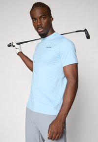 Hellblaues kurzärmliges Golfshirt aus feuchtigkeitsableitendem Material, mit hohem Kragen und dezentem Calvin Klein Logo auf der Brust.
