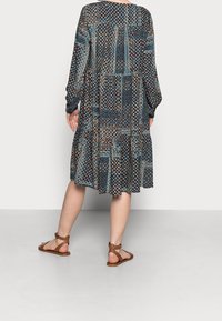 Šaty s květinovým vzorem, dlouhými rukávy, se stupňovitým lemem a v modré, hnědé a béžové barvě. Jsou nošeny s hnědými sandály na pásech.