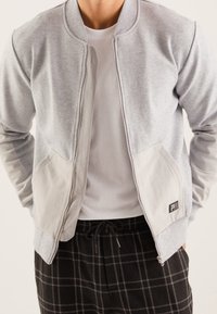 Chaqueta sudadera gris con cierre de cremallera, bolsillos en un gris claro contrastante y un pequeño detalle de logo. Llevada sobre una camiseta blanca y pantalones a cuadros.
