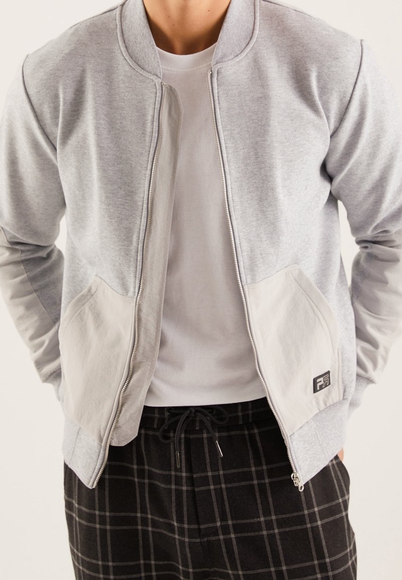 Chaqueta sudadera gris con cierre de cremallera, bolsillos en un gris claro contrastante y un pequeño detalle de logo. Llevada sobre una camiseta blanca y pantalones a cuadros.