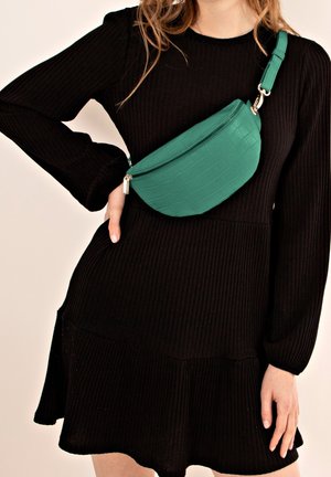 Femme portant une robe noire côtelée à manches longues et un sac bandoulière vert à texture crocodile, fermé par une fermeture éclair, porté en travers de sa poitrine.