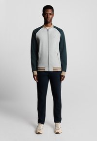 Graue Zip-Jacke mit dunkelgrünen Ärmeln, beigen Akzenten und geripptem Saum. Kombiniert mit marineblauen Jogginghosen und beigen Sneakers.