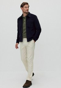 Marinblå kordjacka med bröstficka, grön polo-tröja, krämfärgade byxor och bruna snörskor. Slät textur och strukturerad passform.