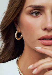 Guldörhängen med en slät yta och lätt vridning, tillsammans med ett guldhalsband. Modellen har långt hår och neutral sminkning.