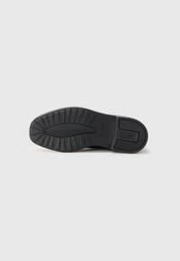 Suela de zapato de goma negra con un patrón texturizado, que presenta un dibujo definido y una marca en el lateral. Forma plana, diseño robusto y material flexible.