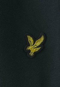 Tessuto nero con una trama liscia, decorato con un logo di aquila gialla ricamato con contorni neri. Il design è preciso e dettagliato.