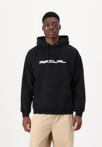 Hanorac negru cu un buzunar frontal și logo alb. Dispune de o glugă cu șnur și manșete ribbate. Purtați cu pantaloni bej.
