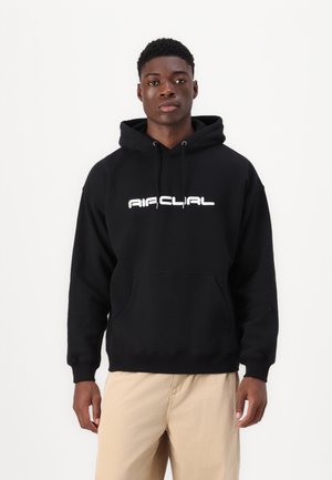 Hoodie noir avec une poche avant et un logo en texte blanc. Comprend une capuche avec cordon de serrage et des poignets côtelés. Porté avec un pantalon beige.